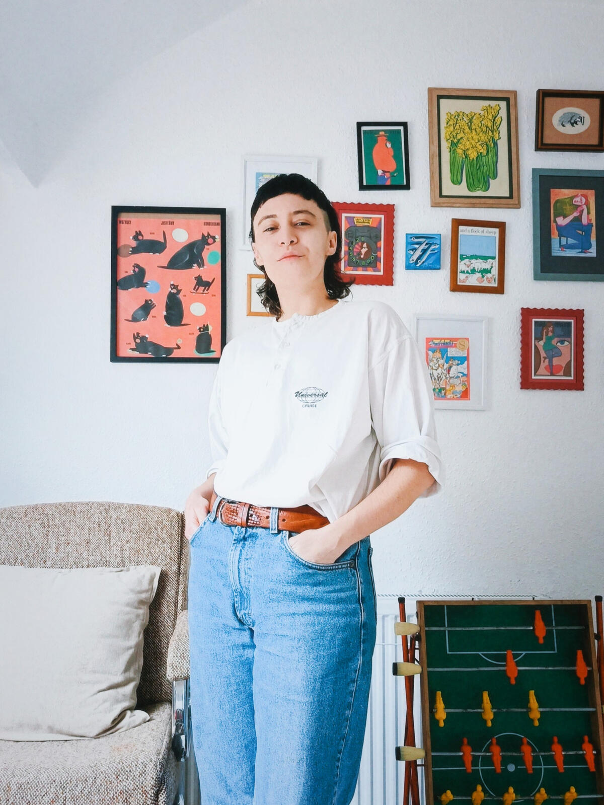 photo of Evie, who is wearing a white t shirt with midlength sleeves, light blue jeans and a brown belt. They have their hands in pockets and are looking into the camera smiling slightly. Evangeline has dark hair in a mullet style, behind them is a wall wi