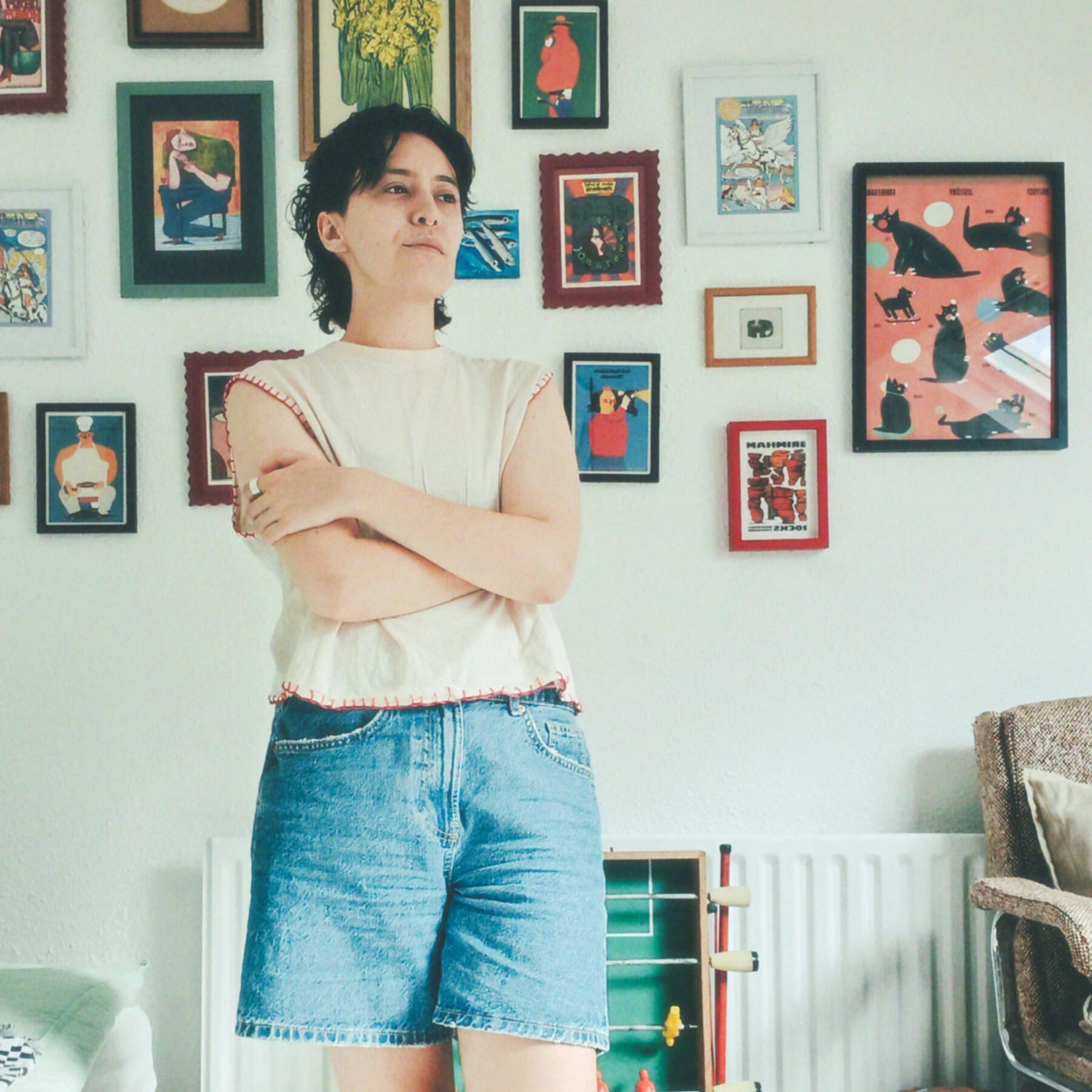 photo of Evangeline with their arms crossed, wearing denim shorts and a cream sleeveless tee with red stitching. Evangeline has dark hair in a mullet style, behind them is a wall with lots of colourful art prints framed upon.